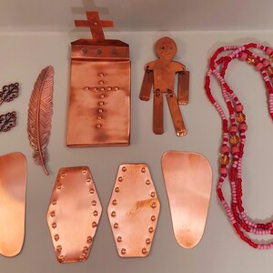 Puede incluir: Una colección de suministros de artesanía de metal color cobre, que incluye llaves, una pluma, una pieza en forma de cruz, una figura con forma de persona y varias formas geométricas. También hay un collar de cuentas con cuentas rojas y rosas.