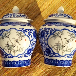 May include: Two matching blue and white ceramic jars with lids. Each jar features a central panel with a bird perched on a bamboo branch, surrounded by a blue floral and scroll design. The lids have a similar pattern.