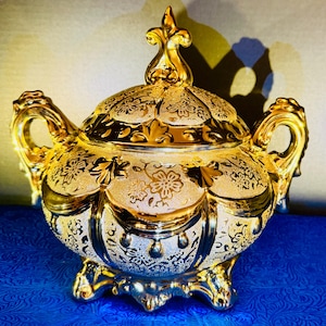 May include: A gold-toned lidded sugar bowl featuring a textured surface with embossed floral patterns, two ornate side handles, and a decorative finial on the lid. The bowl stands on four small feet and has a reflective, metallic finish throughout.