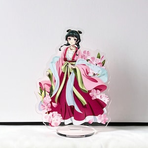Puede incluir: Un expositor acr&iacute;lico transparente con un personaje femenino de anime con un vestido tradicional rosa y rojo, adornado con flores rosas. El personaje tiene cabello negro y est&aacute; sobre una base circular transparente.
