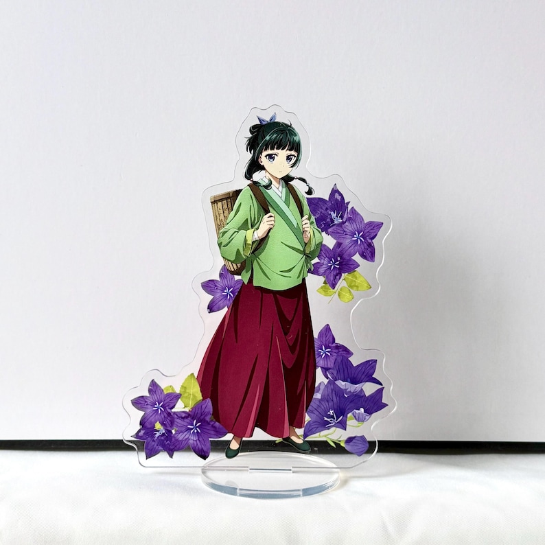 Puede incluir: Un expositor acr&iacute;lico transparente con un personaje de anime femenino con una parte superior verde y una falda burdeos, que lleva una cesta. El expositor est&aacute; decorado con acentos de flores moradas y se apoya en una base transparente.