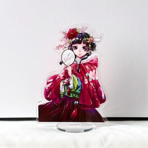 Puede incluir: Figura de pie de acr&iacute;lico transparente de una mujer con un atuendo tradicional rojo y estampado floral. Sostiene un peque&ntilde;o abanico y lleva una corona de flores. La figura tiene una base redonda.