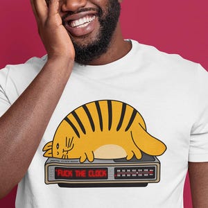 May include: White t-shirt featuring a cartoon cat with orange stripes lying on a vintage radio. The radio displays the text "FUCK THE CLOCK". The person in the image is wearing a yellow beanie.