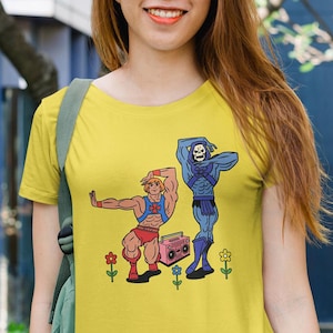 May include: Yellow t-shirt featuring a cartoon illustration of two characters, one muscular and one skeletal, dancing near a pink boombox. The shirt is worn by a person wearing sunglasses and denim shorts.