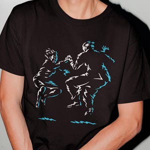 May include: A black short-sleeved t-shirt featuring a graphic print of two figures in motion. The design is rendered in white and light blue outlines, depicting the silhouettes of two people jumping or dancing against the solid black fabric.