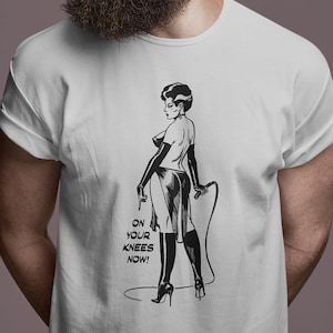 Może przedstawiać: Biała koszulka z czarno-białą ilustracją kobiety w stroju dominatorki, trzymającej bat. Tekst "ON YOUR KNEES NOW!" znajduje się pod ilustracją. Koszulka jest noszona przez osobę w okularach przeciwsłonecznych.