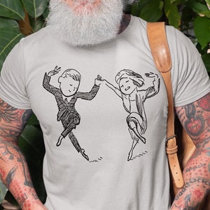 May include: A gray t-shirt with a black and white illustration of a man and woman dancing. The man is wearing a suit and the woman is wearing a dress. The t-shirt is being worn by a person with a white beard and tattoos.