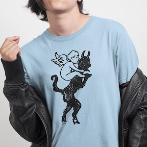 May include: A light blue short-sleeved t-shirt featuring a black graphic print of a winged cherub riding on the back of a horned creature. The shirt is worn under a black leather jacket with ribbed cuffs and is paired with black pants.