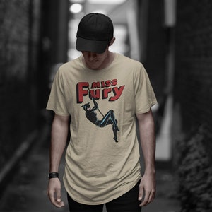 Puede incluir: Una camiseta beige con una ilustraci&oacute;n de c&oacute;mic vintage de una mujer con un traje de gato negro. El texto "Miss Fury" est&aacute; impreso en letras rojas sobre la ilustraci&oacute;n.