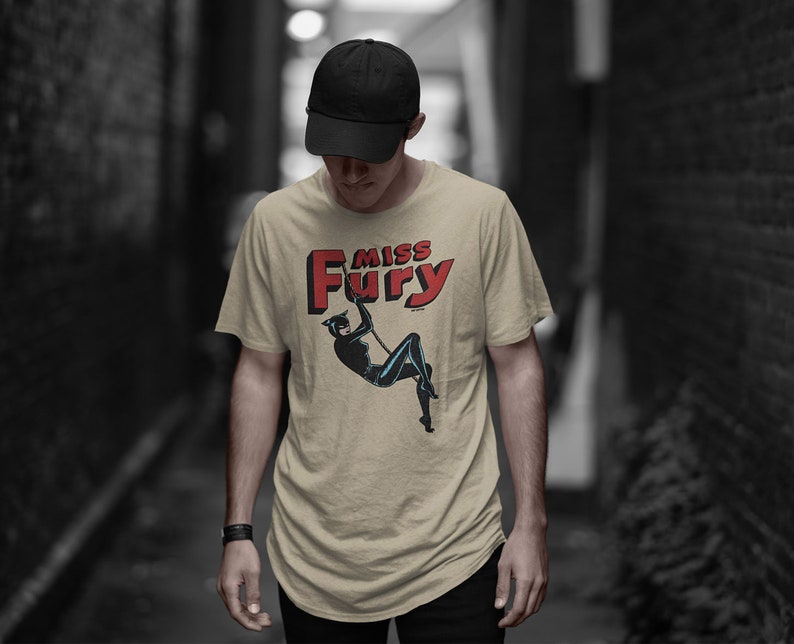 Puede incluir: Una camiseta beige con una ilustraci&oacute;n de c&oacute;mic vintage de una mujer con un traje de gato negro. El texto "Miss Fury" est&aacute; impreso en letras rojas sobre la ilustraci&oacute;n.