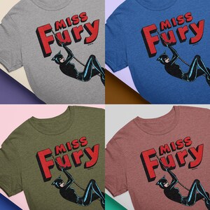 Puede incluir: Cuatro camisetas de diferentes colores con un gr&aacute;fico de estilo c&oacute;mic vintage de una mujer con un traje de gato negro balance&aacute;ndose en una cuerda. El texto "Miss Fury" est&aacute; en letras rojas sobre la mujer.