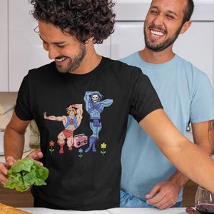 Pu&ograve; includere: T-shirt nero con una grafica a cartoni animati di due personaggi muscolosi, uno in rosso e l'altro in blu, con un boombox rosa e fiori. Due persone sorridono sullo sfondo. Una pagnotta e verdure sono in primo piano.