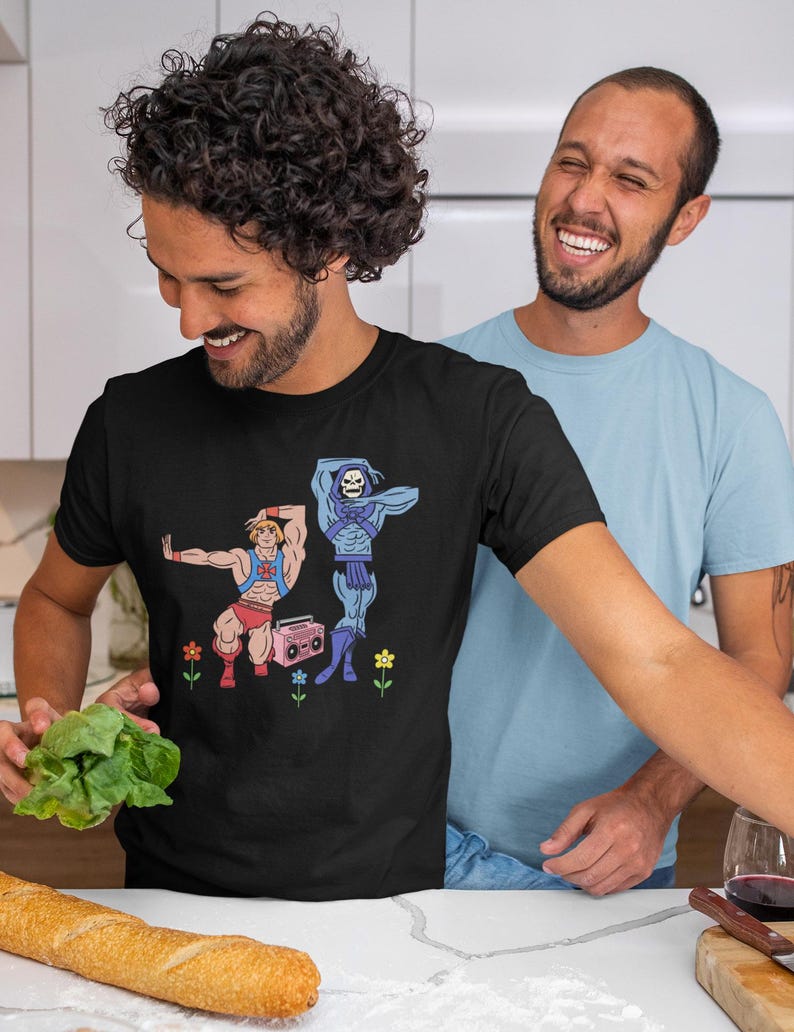 Pu&ograve; includere: T-shirt nero con una grafica a cartoni animati di due personaggi muscolosi, uno in rosso e l'altro in blu, con un boombox rosa e fiori. Due persone sorridono sullo sfondo. Una pagnotta e verdure sono in primo piano.