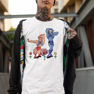 Pu&ograve; includere: T-shirt bianco con un'illustrazione a cartoni animati di due personaggi, uno biondo e uno blu, che posano con un boombox rosa e dei fiori. I personaggi indossano abiti rossi e blu. La maglietta &egrave; indossata da una persona con un berretto blu.