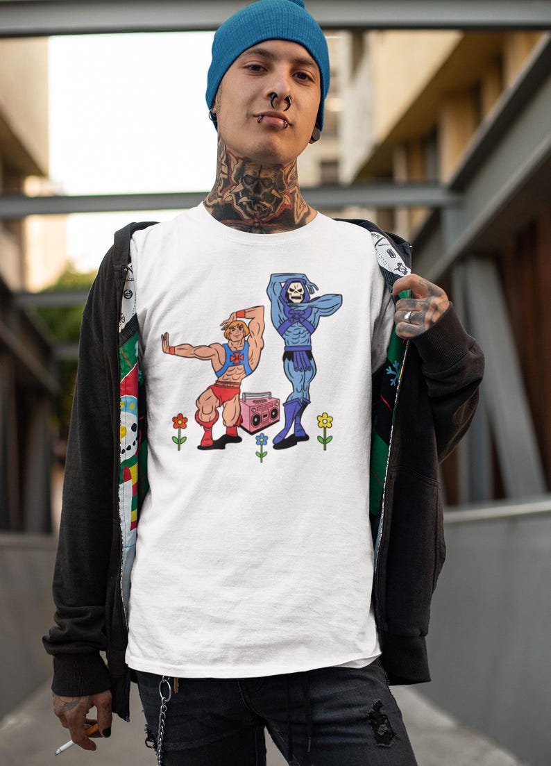 Pu&ograve; includere: T-shirt bianco con un'illustrazione a cartoni animati di due personaggi, uno biondo e uno blu, che posano con un boombox rosa e dei fiori. I personaggi indossano abiti rossi e blu. La maglietta &egrave; indossata da una persona con un berretto blu.