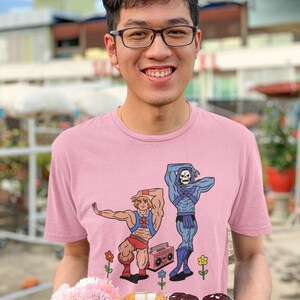 Pu&ograve; includere: Una maglietta rosa chiaro con una grafica a cartoni animati di due personaggi, uno con pantaloncini rossi e l'altro in blu, con un boombox. La persona tiene in mano un vassoio di legno con pasticcini e fiori.