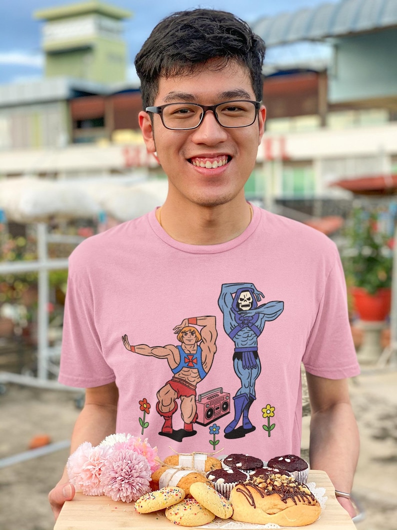 Pu&ograve; includere: Una maglietta rosa chiaro con una grafica a cartoni animati di due personaggi, uno con pantaloncini rossi e l'altro in blu, con un boombox. La persona tiene in mano un vassoio di legno con pasticcini e fiori.
