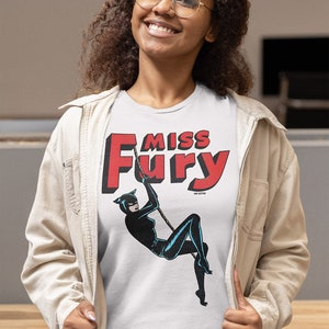 Puede incluir: Una camiseta blanca con un gr&aacute;fico rojo y negro de una mujer con un traje de gato colgando de una cuerda. El gr&aacute;fico dice "Miss Fury" en letras rojas.