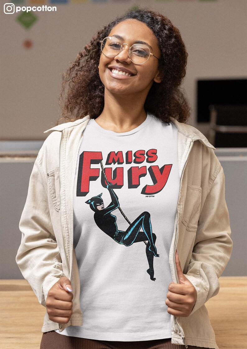 Puede incluir: Una camiseta blanca con un gr&aacute;fico rojo y negro de una mujer con un traje de gato colgando de una cuerda. El gr&aacute;fico dice "Miss Fury" en letras rojas.