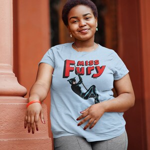 Puede incluir: Una camiseta azul claro con un gr&aacute;fico rojo y blanco de una mujer con un traje de gato con el texto "Miss Fury".