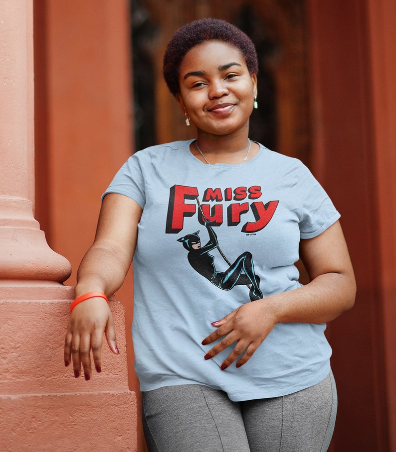 Puede incluir: Una camiseta azul claro con un gr&aacute;fico rojo y blanco de una mujer con un traje de gato con el texto "Miss Fury".