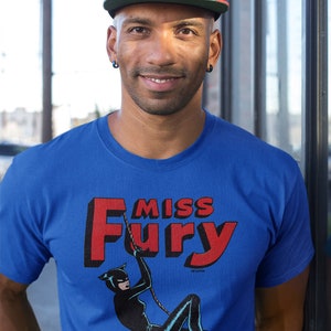 Puede incluir: Una camiseta azul con un gr&aacute;fico rojo y negro de una mujer con un traje de gato colgada de una cuerda. El gr&aacute;fico dice "Miss Fury" en letras rojas.