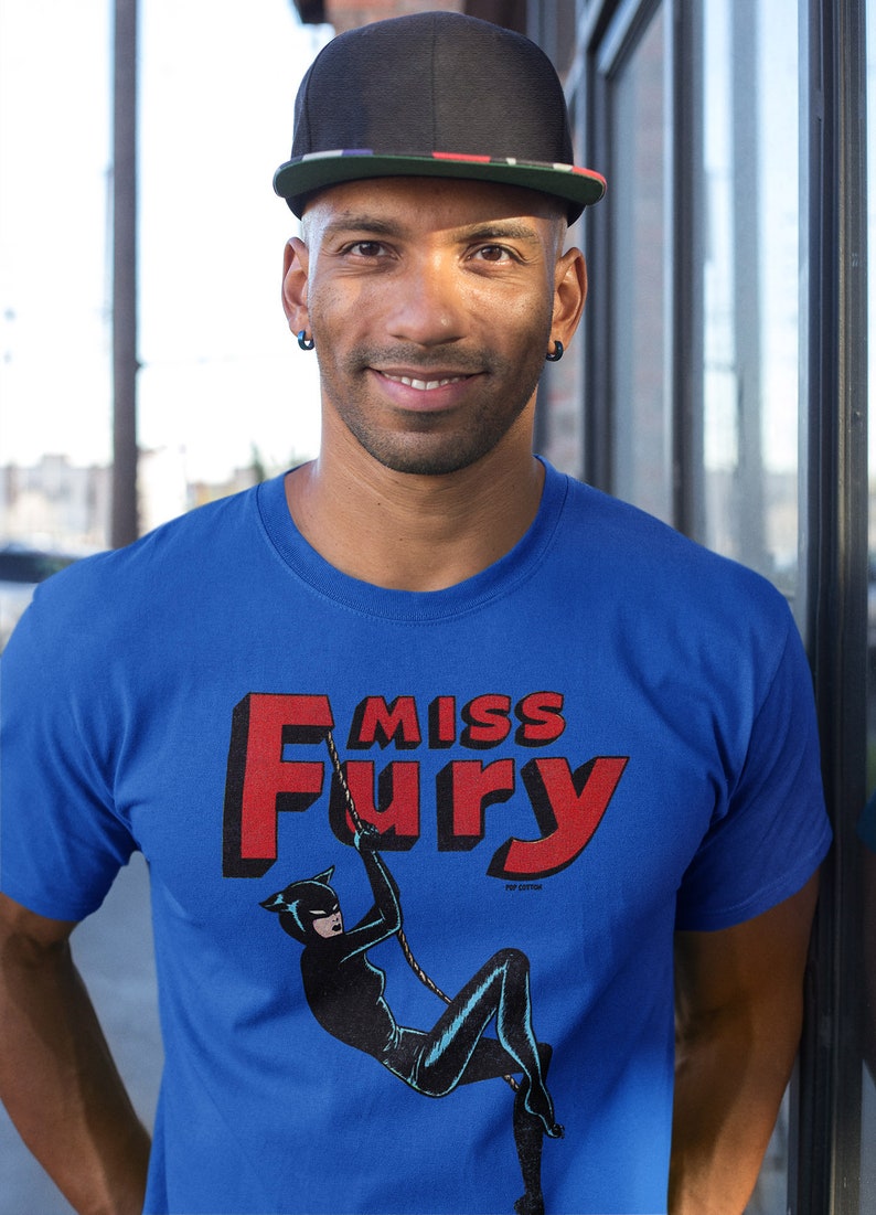 Puede incluir: Una camiseta azul con un gr&aacute;fico rojo y negro de una mujer con un traje de gato colgada de una cuerda. El gr&aacute;fico dice "Miss Fury" en letras rojas.