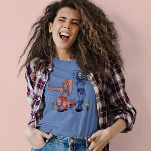 Pu&ograve; includere: Una maglietta blu con un'illustrazione a cartoni animati di due personaggi, uno muscoloso e l'altro scheletrico, che ballano vicino a un boombox rosa e fiori. La maglietta &egrave; indossata da una persona con i capelli ricci e una camicia a quadri.