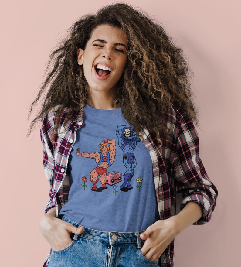 Pu&ograve; includere: Una maglietta blu con un'illustrazione a cartoni animati di due personaggi, uno muscoloso e l'altro scheletrico, che ballano vicino a un boombox rosa e fiori. La maglietta &egrave; indossata da una persona con i capelli ricci e una camicia a quadri.