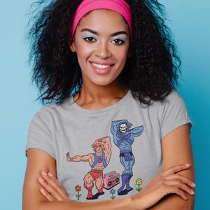 Pu&ograve; includere: Una maglietta grigia con una grafica a cartoni animati di due personaggi, uno con i capelli biondi e l'altro con la faccia da teschio. I personaggi sono in posa con un boombox rosa e dei fiori. La persona nell'immagine indossa una fascia rosa.