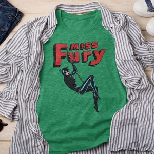 Puede incluir: Una camiseta verde con una ilustraci&oacute;n en blanco y negro de una mujer con un traje de gato balance&aacute;ndose en una cuerda. El texto "Miss Fury" est&aacute; impreso en letras rojas sobre la ilustraci&oacute;n.