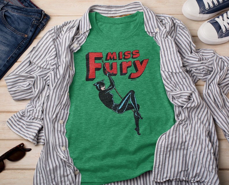 Puede incluir: Una camiseta verde con una ilustraci&oacute;n en blanco y negro de una mujer con un traje de gato balance&aacute;ndose en una cuerda. El texto "Miss Fury" est&aacute; impreso en letras rojas sobre la ilustraci&oacute;n.