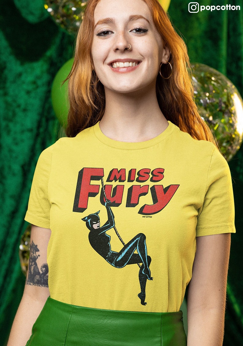 Puede incluir: Camiseta amarilla con un gr&aacute;fico rojo de "Miss Fury" que presenta a una mujer con un traje de gato negro balance&aacute;ndose de una cuerda. El gr&aacute;fico tambi&eacute;n incluye el texto "Pop Cotton".