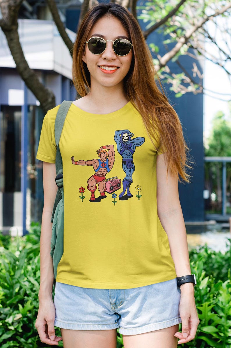 May include: Yellow t-shirt featuring a cartoon illustration of two characters, one muscular and one skeletal, dancing near a pink boombox. The shirt is worn by a person wearing sunglasses and denim shorts.