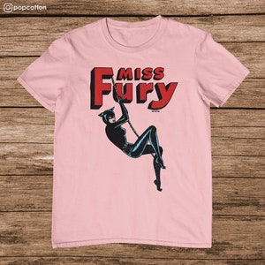 Puede incluir: Camiseta rosa con un gr&aacute;fico de una mujer con un traje de gato negro colgando de una cuerda. El texto "Miss Fury" est&aacute; en letras rojas sobre la mujer.