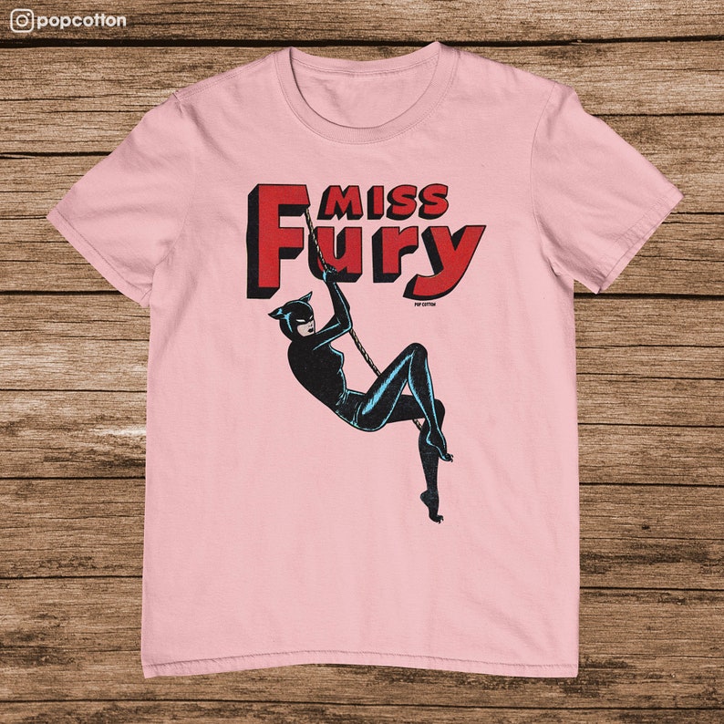 Puede incluir: Camiseta rosa con un gr&aacute;fico de una mujer con un traje de gato negro colgando de una cuerda. El texto "Miss Fury" est&aacute; en letras rojas sobre la mujer.