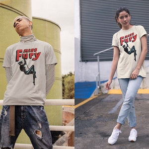 Puede incluir: Una camiseta gris con un gr&aacute;fico rojo y blanco que dice "Miss Fury" y presenta a una mujer con un traje de gato negro. La mujer est&aacute; columpi&aacute;ndose de una cuerda.