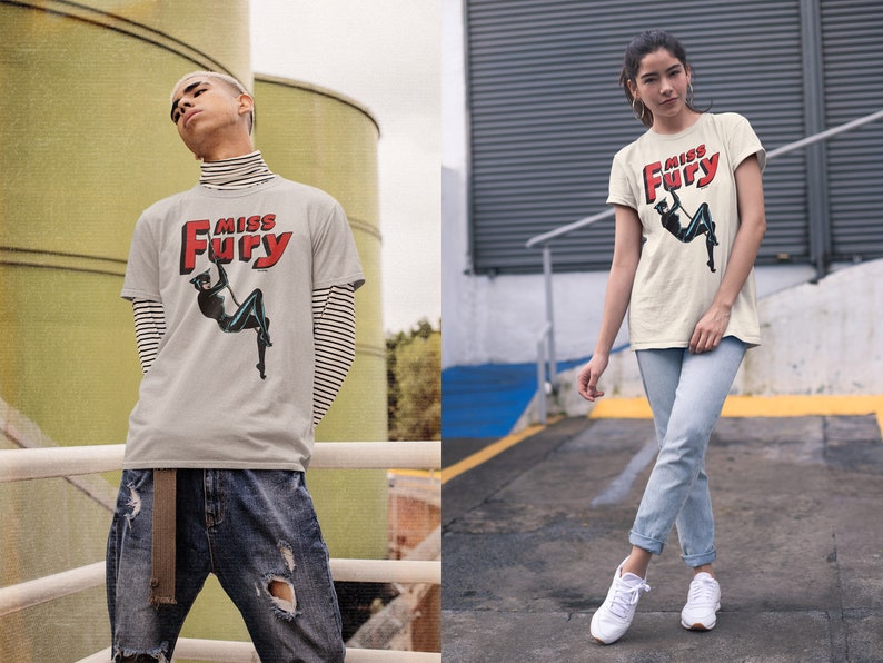 Puede incluir: Una camiseta gris con un gr&aacute;fico rojo y blanco que dice "Miss Fury" y presenta a una mujer con un traje de gato negro. La mujer est&aacute; columpi&aacute;ndose de una cuerda.