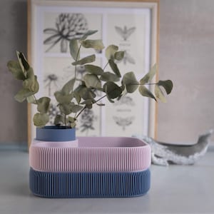 May include: A stacked, ribbed, rectangular container set in blue and pink, holding a small blue vase with green foliage. A framed botanical print and a whale figurine are in the background.