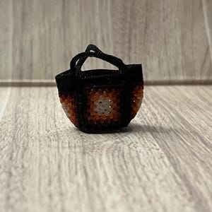 May include: A small, black beaded handbag with a square pattern in shades of orange, white, and brown. The bag has a rounded shape and a small handle. The background is a light brown wooden surface.