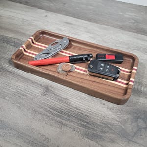 May include: A rectangular wooden tray with a dark brown finish and inlaid stripes of red and light wood. The tray holds a silver folding knife, a red pen, coins, a car key fob, and a black USB drive.