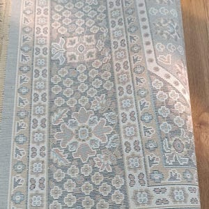 May include: Rolled-up area rug with a detailed floral pattern in shades of beige, blue, and brown. The rug has a fringe on one end and a repeating geometric border design. The rug is made of a woven material.
