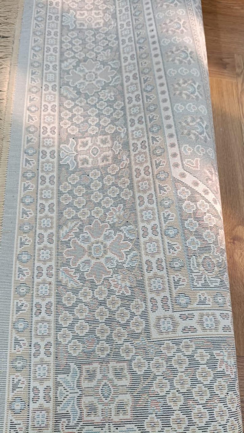 May include: Rolled-up area rug with a detailed floral pattern in shades of beige, blue, and brown. The rug has a fringe on one end and a repeating geometric border design. The rug is made of a woven material.