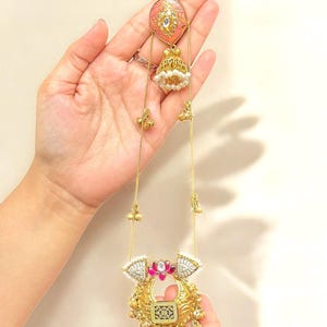 May include: A gold necklace with a pendant featuring a pink and white design, decorated with small gold bells and white pearls. The necklace is held in a hand, highlighting its intricate details and craftsmanship.