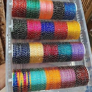 May include: A clear plastic container filled with multiple stacks of colourful bangles. The bangles are in various colours, including red, green, orange, black, blue, pink, yellow, teal, and purple. Each stack is approximately 5 cm in diameter.