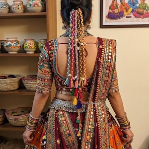 May include: A maroon and orange lehenga choli featuring intricate embroidery, mirror work, and colorful borders. The outfit includes a matching dupatta, a silver waist belt, and stacked bangles. The hair is styled in a bun with colorful braided hair extensions adorned with small metallic coins and white seashells.