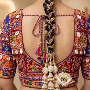 May include: A back view of a blouse featuring intricate embroidery in red, orange, and blue with mirror accents and white cowrie shells. A long, thick braid is adorned with gold-toned metallic trim and a large, multi-tiered white tassel accessory.