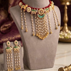 May include: A gold-toned choker necklace and matching drop earrings set featuring green and red enamel elephant motifs, circular accents, and dangling white pearl-like beads with small gold-toned metal spheres at the ends of the chains.