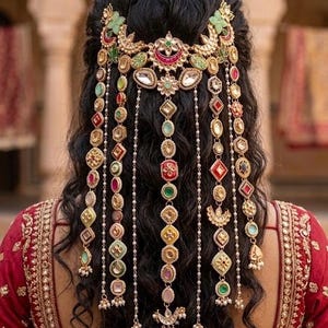 May include: A gold-toned hair accessory featuring intricate metalwork, colorful enamel accents in red, green, and yellow, and dangling chains adorned with geometric shapes and small white beads, worn over dark, wavy hair with a red embroidered garment.