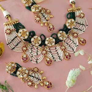 May include: An ornate necklace and earring set. The necklace features multiple strands of white beads, interspersed with green beads, and gold-toned floral accents with red and clear stones. The earrings mirror the necklace design, with teardrop-shaped pendants.